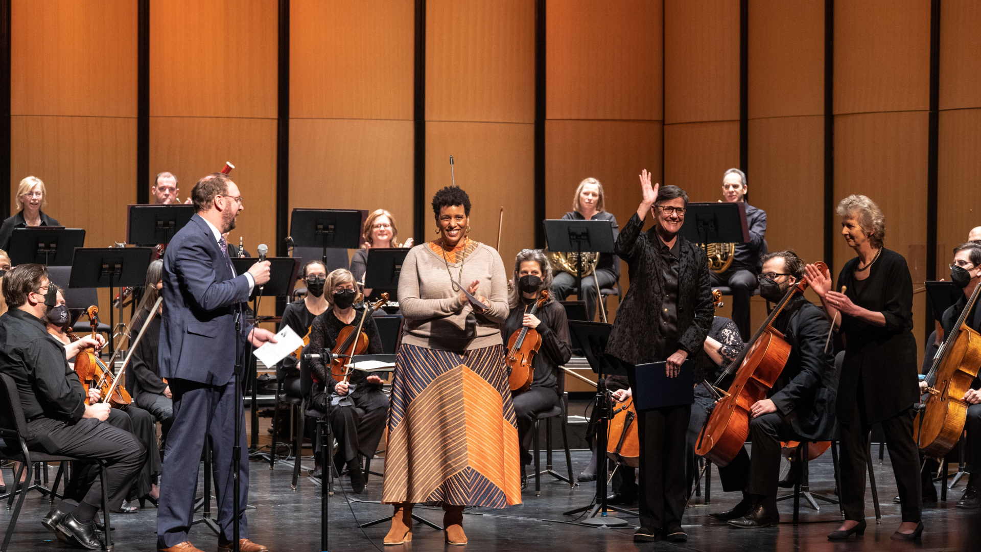 Declan McGovern, Erin Harkey, Amy Eschleman, and Dame Jane Glover wave and clap on a stage surrounded by the MOB Orchestra