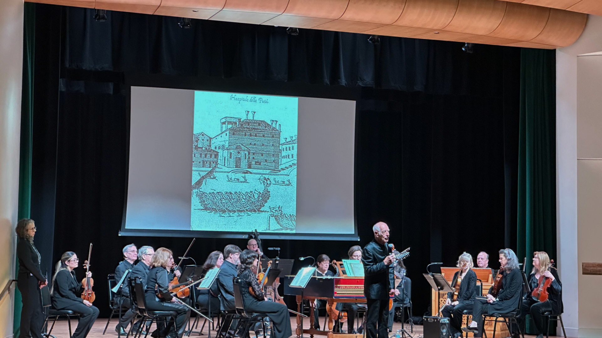 Nicholas Kraemer stands in front of the MOB Orchestra and a slideshow